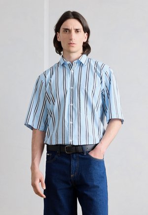 Junger Mann trägt ein hellblaues kurzärmeliges Hemd mit schwarz-weißen vertikalen Streifen und dunkelblaue Jeans, steht mit einer Hand in der Tasche.