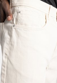 O mână de culoare închisă odihnindu-se în buzunarul din față al unei perechi de pantaloni din denim off-white, cu cusături vizibile și nituri metalice.