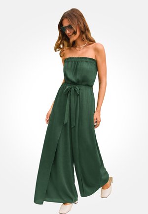 Kobieta ubrana w ciemnozielony, bezramienny kombinezon z szerokimi nogawkami, z paskiem przewiązanym w talii, białe buty, okulary przeciwsłoneczne i warstwowe złote naszyjniki.