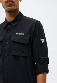 Άνδρας που φοράει μαύρο πουκάμισο Columbia Titanium με κουμπιά, τσέπες στο στήθος και μανίκια που είναι γυρισμένα, εμφανίζοντας το λογότυπο και το κείμενο "Omni-Shade" στο μανίκι.