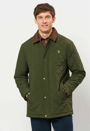 Homme portant une veste matelassée verte avec un col marron, les mains dans les poches, souriant et debout devant un fond clair uni.