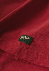 Czerwona bawełniana tkanina o gładkiej teksturze, z zieloną metką "Superdry" uszytą na brzegu. Prosty design, bez wzorów ani dodatkowych detalów.