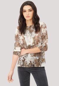 Blusa marrón de estampado de serpiente con acentos florales, de mangas largas arremangadas hasta el codo y un corte relajado, combinada con jeans gris oscuro.