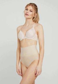 Kvinne iført beige høy midje shaping-truser og matchende lett polstret BH, stående mot en ensfarget lys bakgrunn.