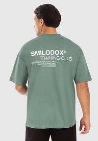 Grøn bomulds t-shirt med korte ærmer, en afslappet pasform og fed hvid tekst på ryggen, der inkluderer brandnavn og motiverende sætninger.
