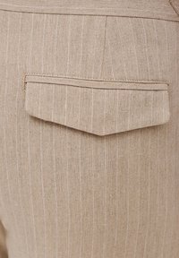 Beżowa tkanina lniana z cienkimi białymi paskami, z kwadratową tylną kieszenią z wyszywaną bordiurą. Brak widocznych elementów metalowych lub ozdób.