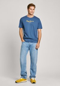 Zila kokvilnas T krekls ar dzelteno "Pepe Jeans London" logotipu, pārī ar gaiši ziliem džinsiem un dzeltenām sporta kedām. Ikdienas piegriezums un īsām piedurknēm.