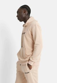 Sudadera beige hecha de tejido suave, con un bolsillo delantero, capucha con cordón y texto de logo en negro. Combinada con pantalones de chándal a juego.