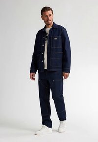 Dunkelblaue Jeansjacke mit Fronttaschen, kombiniert mit passenden Hosen. Das Modell trägt ein helles T-Shirt und weiße Sneaker.