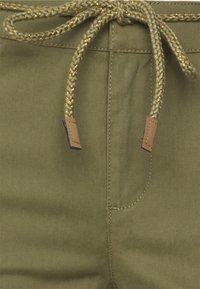 I pantaloni verde oliva presentano un cordino intrecciato oro e verde con dettagli in pelle marrone. Tessuto liscio con tasche cucite e cuciture.