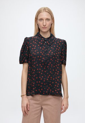 Femme aux cheveux blonds lisses portant un chemisier noir à motifs floraux rouges et un pantalon beige, debout sur un fond blanc uni.