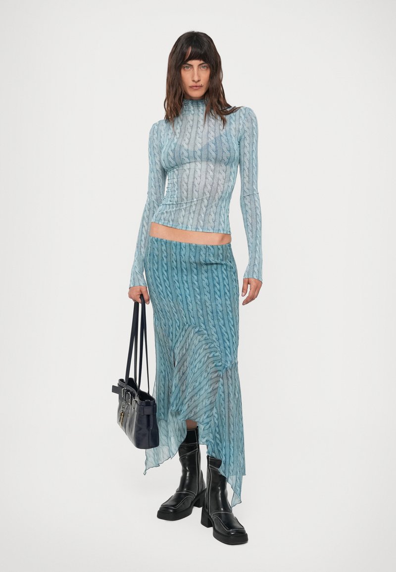 Kvinna med en blåmönstrad långärmad topp och matchande asymmetrisk kjol, som håller en svart handväska och har på sig svarta ankelstövlar.