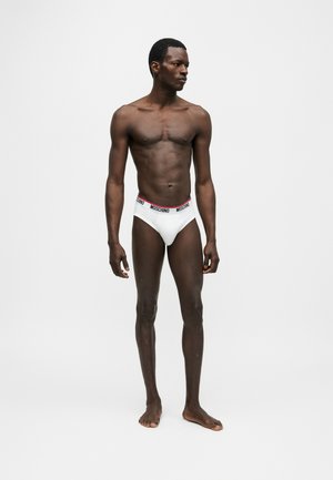 Male model standing barefoot against plain background wearing white Moschino briefs with branded waistband, looking to the side.