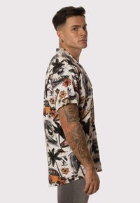 Camisa de manga corta en tejido blanco con estampado tropical colorido, que presenta palmeras, flores y elementos de surf. Corte holgado con escote redondo.