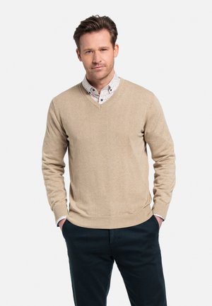 Beige v-hals trui van zachte gebreide stof, gecombineerd met een gestreept overhemd. Het model heeft de handen in de zakken en draagt donkere broeken.