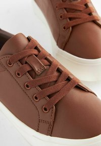 Brune lædersneakers med flade brune snørebånd, rund tå og hvide gummisåler; med syninger og metaløjer.