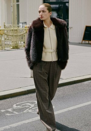 Chaqueta de pelaje sintético marrón sobre un suéter crema, ceñida a la cintura, combinada con pantalones de pata ancha en cuadro marrón. La modelo camina por la calle.