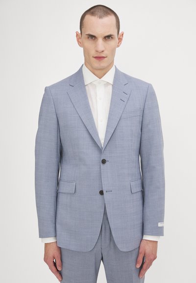 Abito su misura di colore azzurro chiaro con tessuto strutturato. Revers a punta, due bottoni e una tasca sul petto. Design sobrio con una camicia bianca fresca.