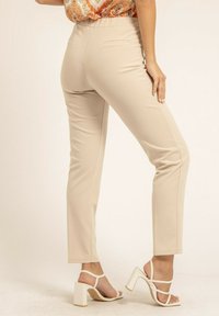 Pantalons beige ajustés avec une coupe slim, texture lisse, taille standard et deux poches arrière. Associés à des sandales blanches à talons carrés.
