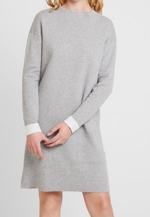 Frau trägt ein langärmeliges, graues Strickkleid mit weißen Manschetten, steht mit sanft verschränkten Händen vor einem schlichten Hintergrund.