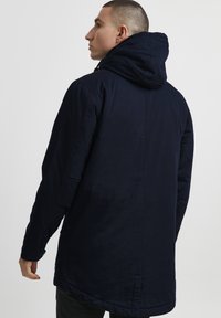 Dark navy parka jacket with a hood, featuring a smooth texture, side pockets, and a long cut that extends past the waist.