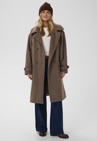 Brązowy wełniany płaszcz z szerokim kołnierzem, dwurzędowymi guzikami, bocznymi kieszeniami, zestawiony z białą bluzką i ciemnoniebieskimi szerokimi spodniami dżinsowymi.