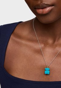 Turkusowy wisiorek w kształcie niedźwiedzia na srebrnym łańcuszku, noszony na ciemnoniebieskim topie. Wisiorek ma miękkie, teksturowane wykończenie.