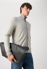 Lys grå zip-genser med ribbede mansjetter, kombinert med en svart skinn clutch-veske med jevn tekstur og avrundede kanter.