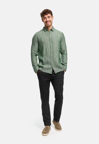 Junger Mann lächelnd, trägt ein grünes Leinenhemd mit Knöpfen, schwarze Hose und beigefarbene Slipper, steht mit den Händen in den Taschen.