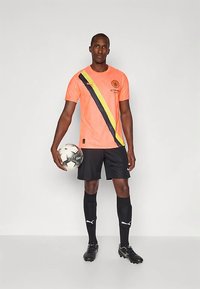 Moški nosi koralno-naravno nogometno majico z rumenimi in črnimi diagonalnimi črtami, črne hlače, nogavice, kopačke ter drži nogometno žogo ob sebi.