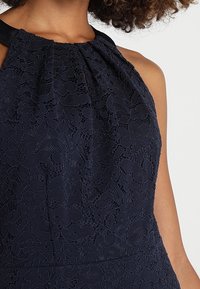 Gros plan d'une robe en dentelle bleu marine sans manches avec des motifs floraux et des plis près de l'encolure ronde, portée par une personne aux cheveux bouclés.