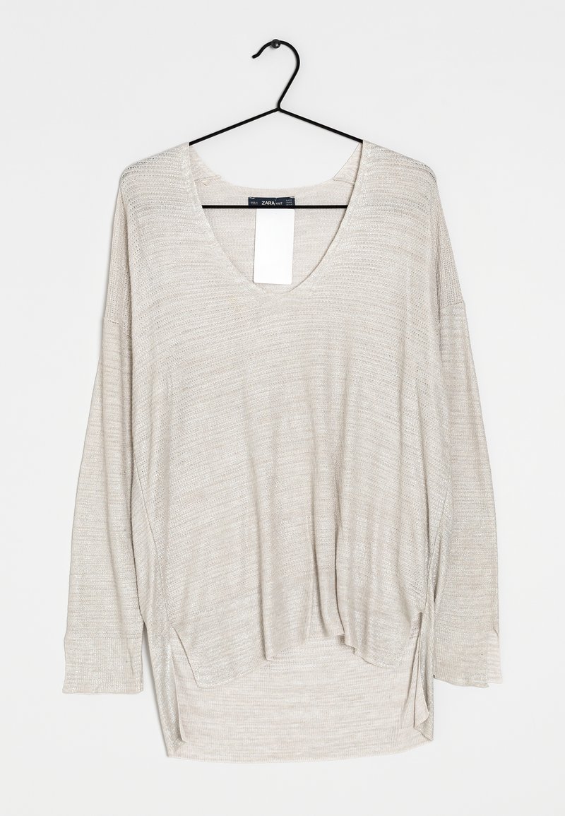 Langarmshirt in hellbeige mit V-Ausschnitt, das ein strukturiertes Strickdesign und Seitenschlitze am Saum aufweist, hängt an einem schwarzen Haken.