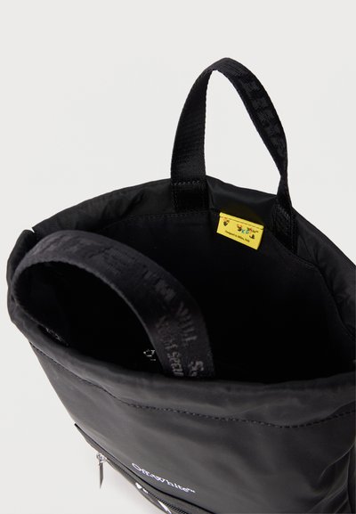 Μαύρη τσάντα tote με υφασμάτινες λαβές και μια μικρή κίτρινη ετικέτα στο εσωτερικό, με μπροστινή τσέπη με φερμουάρ και λογότυπο "Off-White".