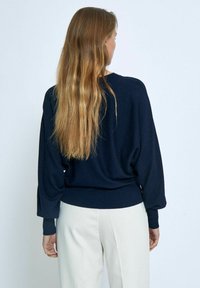 Dunkelblauer Pullover mit langen, lockeren Ärmeln, gerippten Bündchen und Saum. Weiches Strickmaterial, lässige Passform, getragen über hellen Hosen.