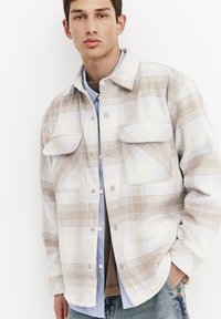 Plaid shirt jacket in beige and light blue tones, featuring snap buttons, two chest pockets, and a relaxed fit. Made of soft, textured fabric.