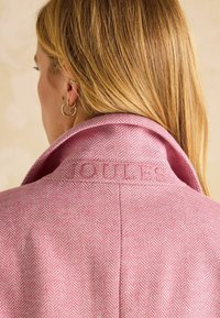 Roosa kalasabamustri jakker, millel on kokkuvolditud krae ja reljeefne bränditekst. Sile kangas, millel on pehme tekstuur.