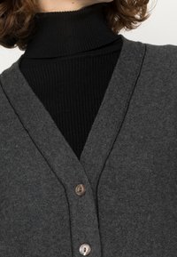 Grå cardigan med V-ringning, tillverkad av strukturerat tyg. Har två knappar och är lagd över en svart ribbad polotröja.
