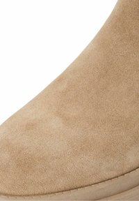 Gros plan sur une bottine en daim beige avec un bout arrondi et un détail de semelle surpiquée.