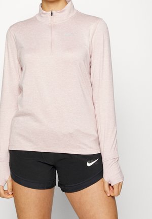 Femme portant un haut zippé à manches longues rose clair et un short de sport noir Nike sur fond uni.