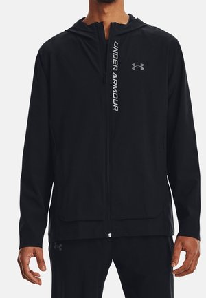 Mand iført sort Under Armour hættetrøje med lynlås og hætte, logo på brystet og teksten "UNDER ARMOUR" langs lynlåsen foran.