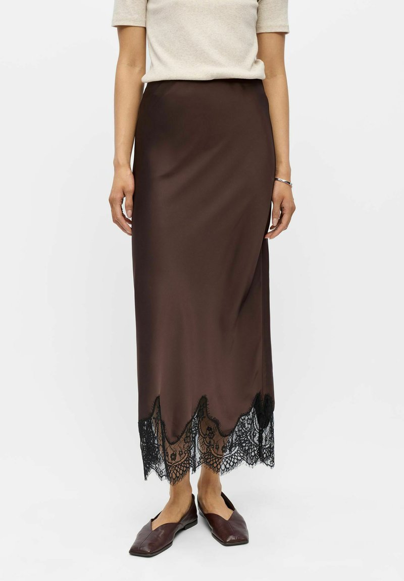 Femme portant une jupe midi en satin marron avec bordure en dentelle noire, un top beige à manches courtes, des chaussures plates marron et un bracelet argenté à son poignet gauche.