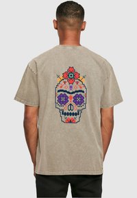 Beige bomull t-skjorte med et fargerikt hodeskalledesign på baksiden, prydet med blomster mønstre i lilla, rød og oransje. Ledig passform.