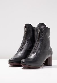 Pár černých texturovaných kožených kotníkových bot s předním zipem a blokovými podpatky na bílém povrchu a s světlým pozadím.