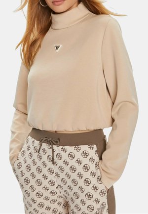 Donna che indossa un top corto beige a maniche lunghe con collo alto e logo, abbinato a pantaloni beige fantasia con coulisse in vita e tasche.