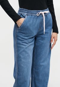 Άτομο που φορά μπλε τζιν παντελόνια φαρδιά με ελαστική μέση και λευκό κορδόνι, μαύρο μακρυμάνικο τοπ, χέρι στην τσέπη.