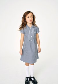 Niebieska i biała sukienka w kratkę z krótkimi rękawami, kołnierzykiem, pięcioma guzikami i kieszonką na piersi; zestawiona z białymi skarpetkami i czarnymi butami.