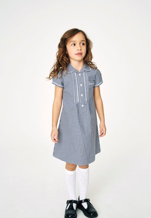 Ruuduinen puuvillamekko sinivalkoisena, jossa on lyhyet hihat, kaulus, viisi nappia ja rintatasku; yhdistetty valkoisiin sukkiin ja mustiin kenkiin.