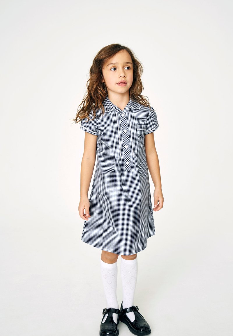 Niebieska i biała sukienka w kratkę z krótkimi rękawami, kołnierzykiem, pięcioma guzikami i kieszonką na piersi; zestawiona z białymi skarpetkami i czarnymi butami.