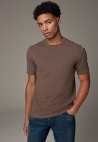 Camiseta corta de manga en color marrón, hecha de algodón, con un escote redondo y un diseño ajustado, combinada con unos jeans de mezclilla azules.
