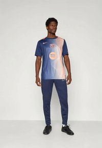 Man staat in een blauw en perzikkleurig FC Barcelona voetbalshirt, marineblauwe Nike broek, witte sokken en zwarte voetbalschoenen tegen een effen achtergrond.